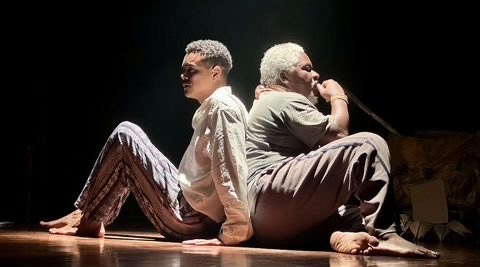 Atores Lincoln Oliveira e Fernando Porto sentados de costas um para o outro no palco durante cena da peça Abdias do Nascimento