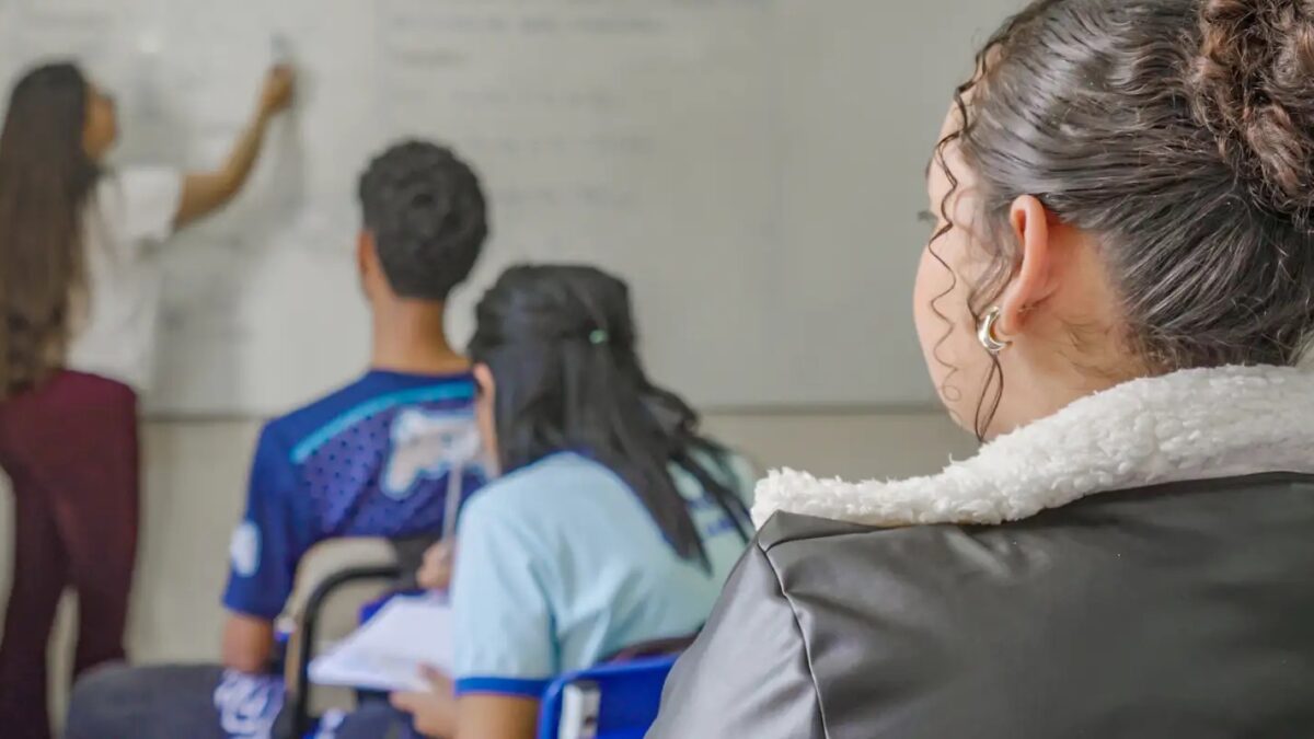 Estudantes assistindo aula em sala com professora escrevendo no quadro branco