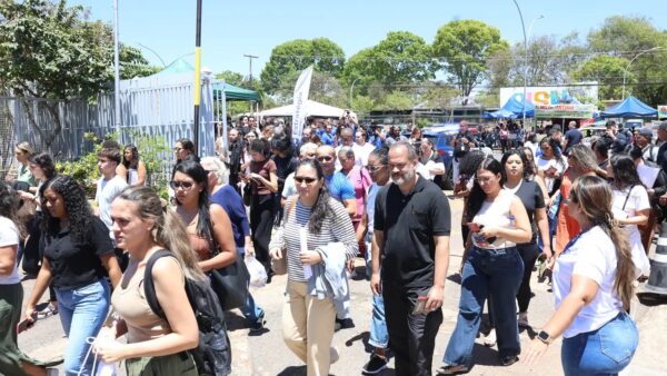 Multidão de candidatos caminhando em fila do lado de fora do portão de um local de prova de concurso público