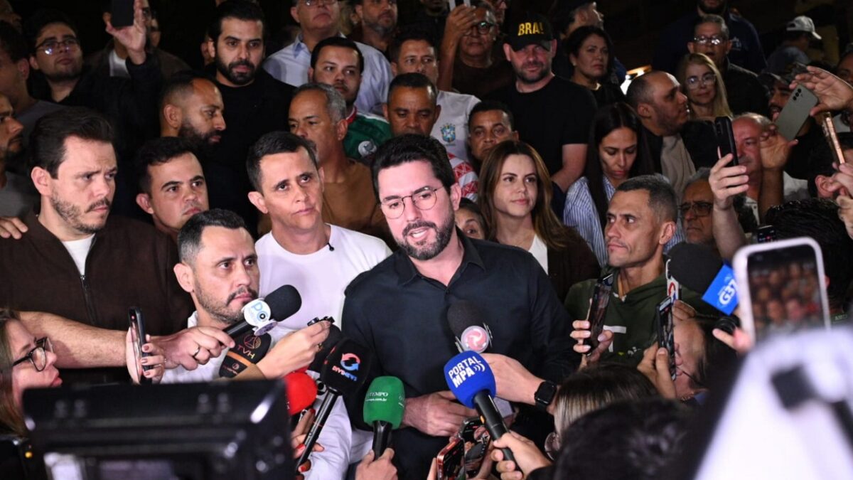 Político fala a jornalistas cercado de apoiadores e microfones durante entrevista coletiva à noite