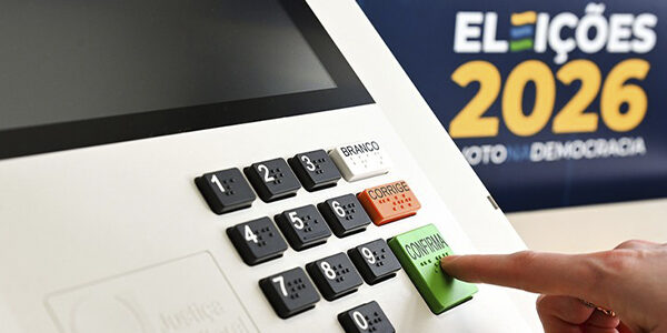 Close do teclado da urna eletrônica com dedo pressionando botão confirma, ao fundo banner Eleições 2026