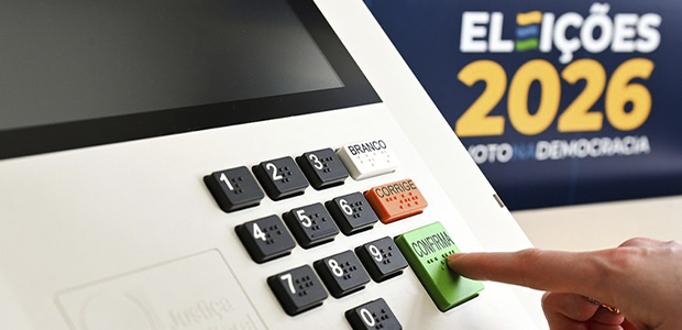 Close do teclado da urna eletrônica com dedo pressionando botão confirma, ao fundo banner Eleições 2026