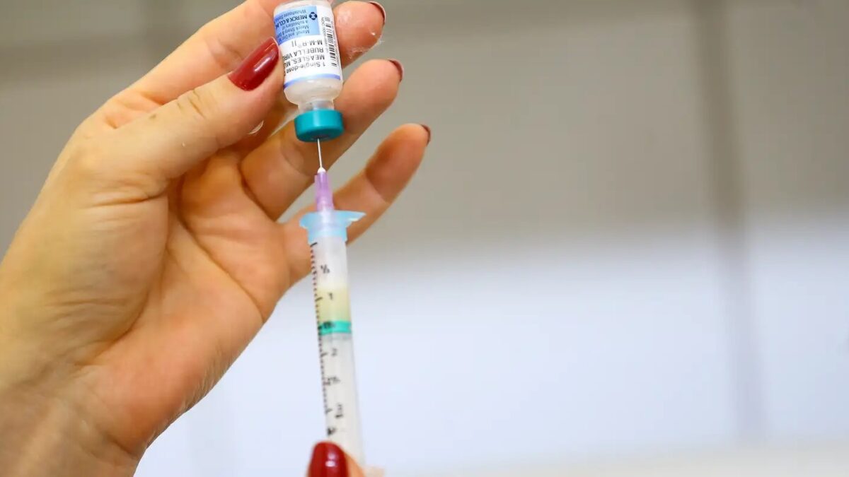 Mão segurando frasco e seringa de vacina contra sarampo, caxumba e rubéola