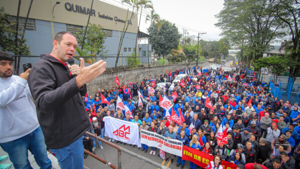 Dirigente sindical discursa ao microfone para multidão de metalúrgicos com bandeiras em frente à fábrica em Diadema