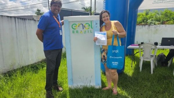 Representante da Enel e moradora sorrindo ao lado de totem da Enel Brasil durante entrega de geladeira nova em Mauá