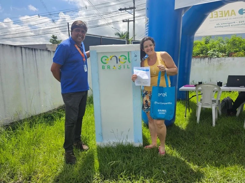 Representante da Enel e moradora sorrindo ao lado de totem da Enel Brasil durante entrega de geladeira nova em Mauá