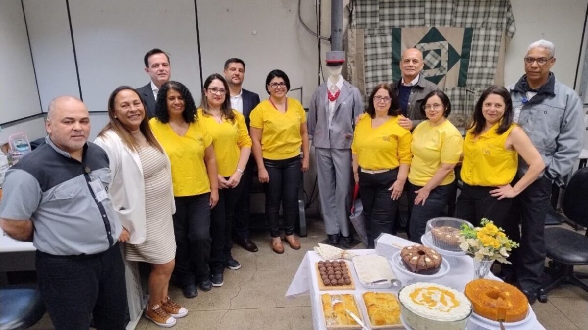 Grupo de pessoas com camisetas amarelas posando ao lado de manequim com uniforme reformado e mesa de doces durante evento do projeto Linha da Moda em Mauá