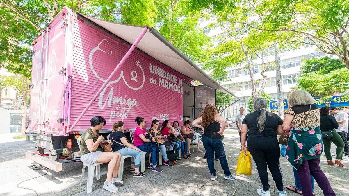 Mulheres sentadas aguardando exame na Carreta da Mamografia, veículo rosa estacionado em área arborizada de São Bernardo do Campo