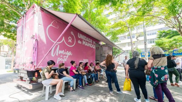 Mulheres sentadas aguardando exame na Carreta da Mamografia, veículo rosa estacionado em área arborizada de São Bernardo do Campo
