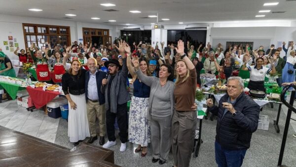 Grupo de voluntários e autoridades acenando em salão decorado com bandeiras e mesas de comida durante preparativos da Festa Italiana de São Caetano