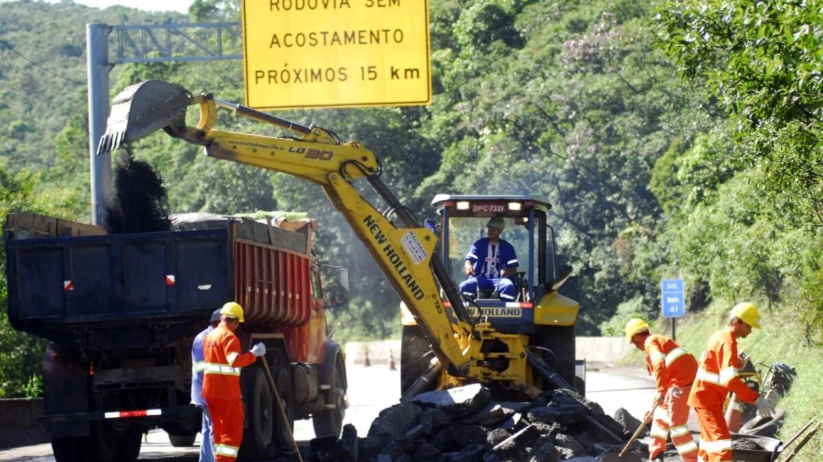 Trabalhadores e escavadeira realizam obras de manutenção em trecho de rodovia com placa Rodovia sem acostamento
