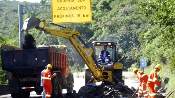 Trabalhadores e escavadeira realizam obras de manutenção em trecho de rodovia com placa Rodovia sem acostamento