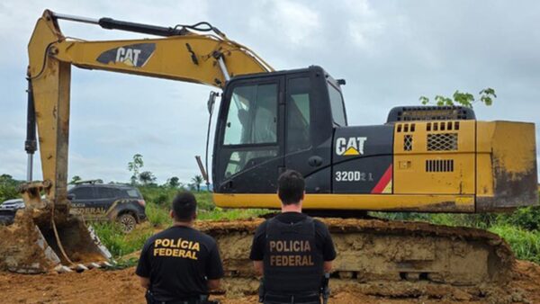 Dois agentes da Polícia Federal de costas diante de uma escavadeira apreendida em garimpo no Pará